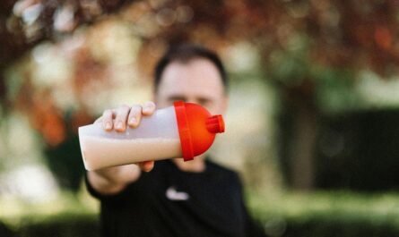 Focus on a man holding a plastic protein shake tumbler outdoors with a blurred background.