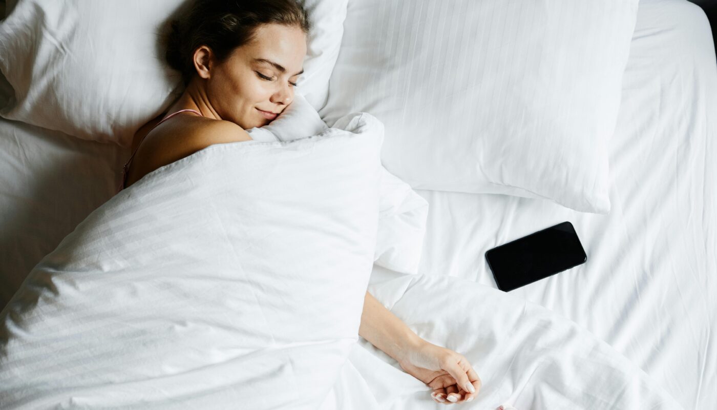 A young woman sleeps comfortably in bed with a smartphone beside her, wrapped in white sheets.